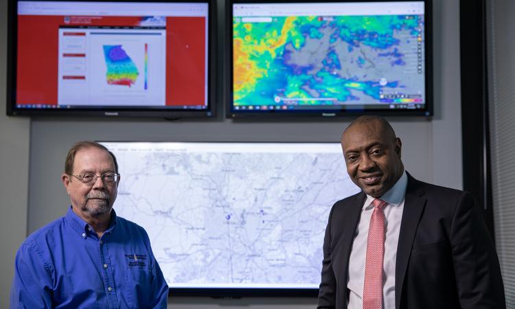Researchers John Trostel (GTRI) in a blue shirt and Marshall Shepherd (University of Georgia) in a black suit and pink tie. Behind them on a wall are three large monitors showing weather maps and radar images. (Credit: Sean McNeil, GTRI)