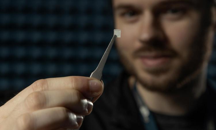 Research Scientist Connor Frost shows a tiny square of materials in a pair of tweezers. The electrical device patterns capture radio frequency energy to alter the properties of the tiny structure. (Photo: Christopher Moore)