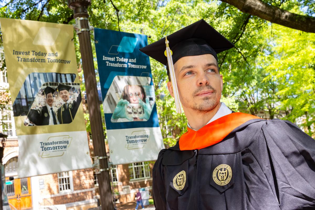 Thomas Vandiver, in master's regalia, stands in front of a campaign banner on campus