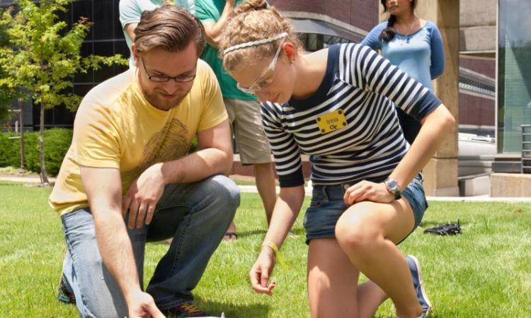 Two students kneel to look at a drone on Tech Lawn with other students in the background