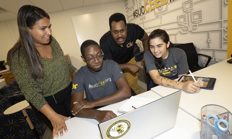 Valentina De La Fe & Cedric Trice working with two students, seated at a table with a laptop, in the CEED office.