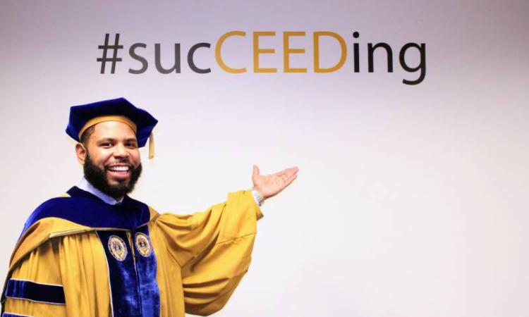 A Ph.D. graduate in tam and gown points to the #sucCEEDing hashtag on a wall.