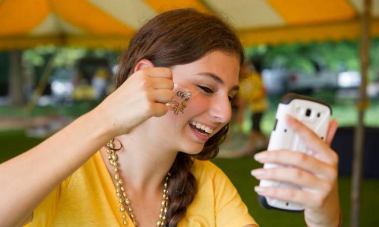 A female student uses her phone as a mirror while she applies a temporary tattoo of Buzz and the interlocking GT logo to her cheek.