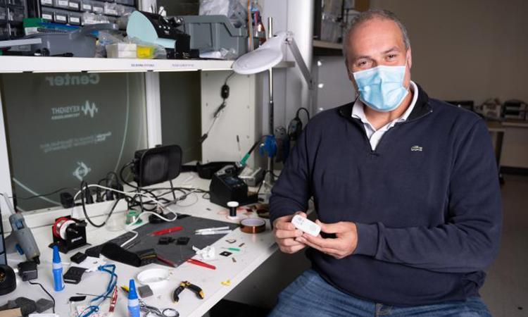 Omer Inan holds a small device at a worktable covered in tools and wires.