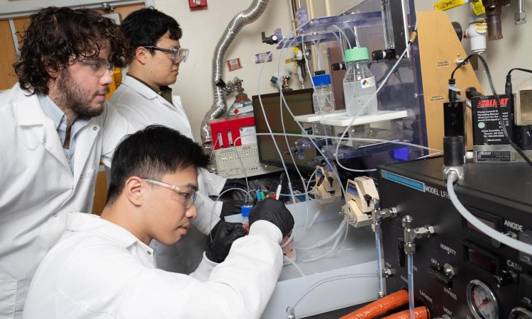 Three men in lab coats working at a bench on an experimental setup with tubes, vials, and pumps.