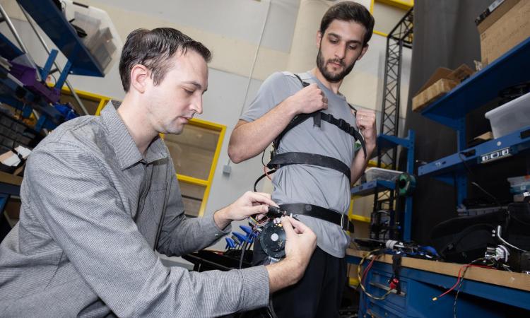 Two people in a lab make adjustments to a robotic exoskeleton