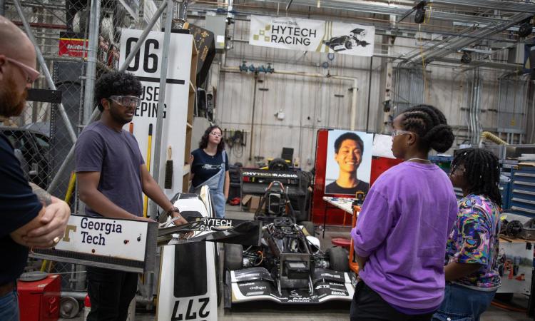 A group of campers learn from current students with a car in the background