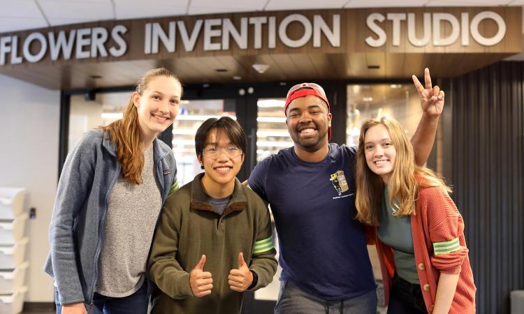 Four students pose arm in arm smiling in front of the Flowers Invention Studio