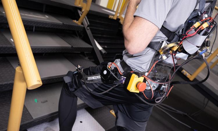 a person wearing a robotic assistive device on his back and legs climbs a set of stairs in a lab