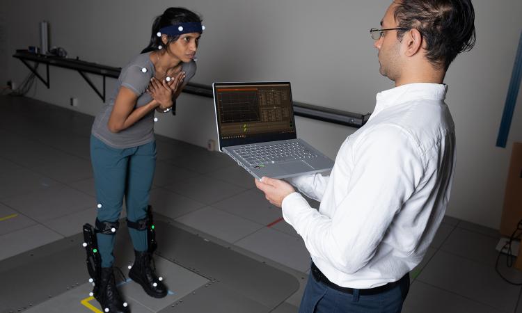 a person wearing biosensors stands on a moving platform while another person tracks movement on a laptop