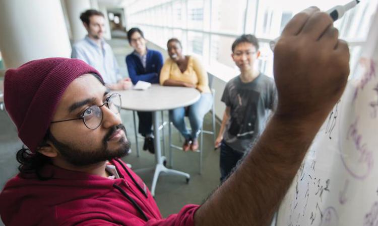 A student writes on a white board while other students look on.