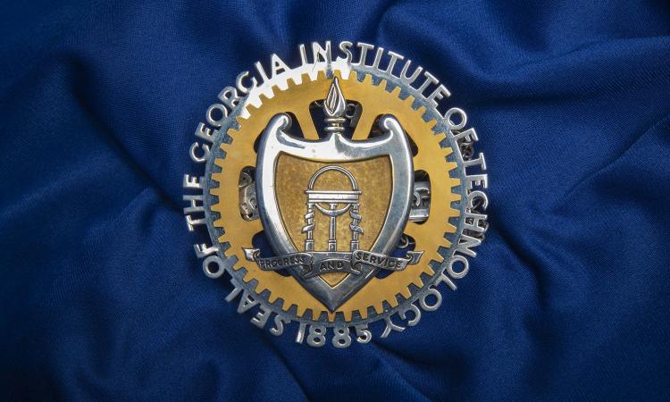 A medal in the shape of the Georgia Tech seal laying on navy blue fabric.