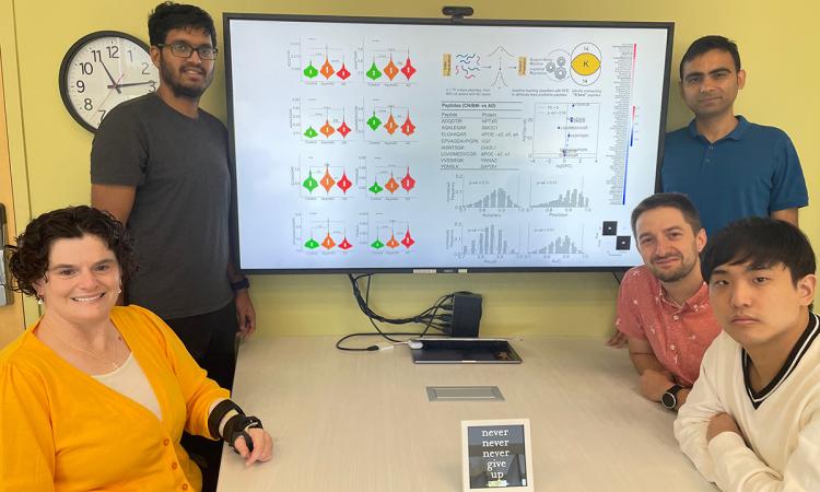 Cassie Mitchell at a table with her Ph.D. students. On the screen above the table is a data slide showing their work on an integrative machine learning algorithm to predict risk of neurodegenerative disease progression based on patient proteins, genotype, demographics, cognitive testing, and medical history. Clockwise from top left are Irfan Al-Hussaini , Raghav Tandon, David Kartchner, Albert Lee, and Mitchell. (Photo Courtesy: Cassie Mitchell)