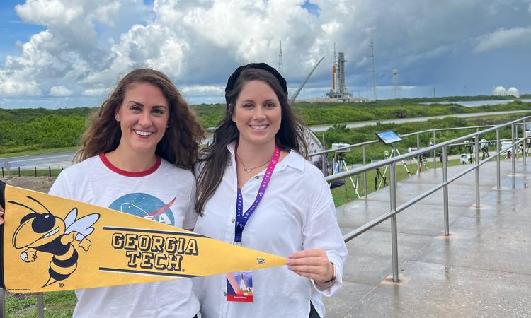 NASA Social attendees Kate Gunderson, MSAE 2018, and AE Communications Manager Kelsey Gulledge with a Georgia Tech pennant and the Artemis I rocket in the background.