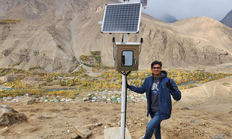 Mirza Samnani stands atop a brown mountain in Tajikistan with one of his avalanche monitoring stations: a pole with solar panel and various weather gauges at the top and a silver metal box below with electronics. Below in the background rooftops from a village are visible.