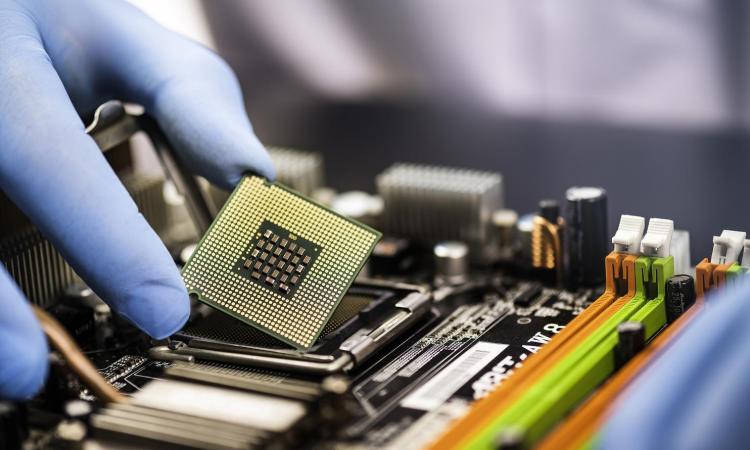 A gloved hand holds a square of silicon with microchips above a board of electronic parts.