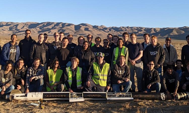 A large group of students poses with their rocket after a successful launch in the desert.