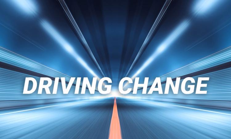 A blurred image of a roadway with streaking lights extending into the distance. Overlaid text reads "Driving Change"