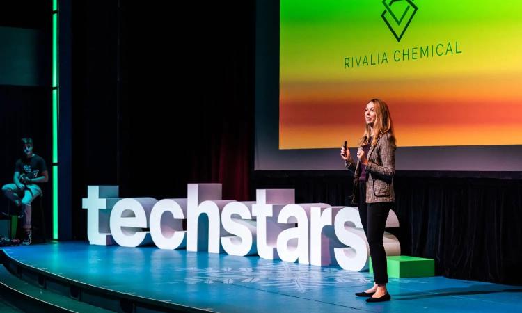 Laura Stoy speaks on stage nex to large letters spelling "techstars" as part of the Techstars Alabama EnergyTech Accelerator.