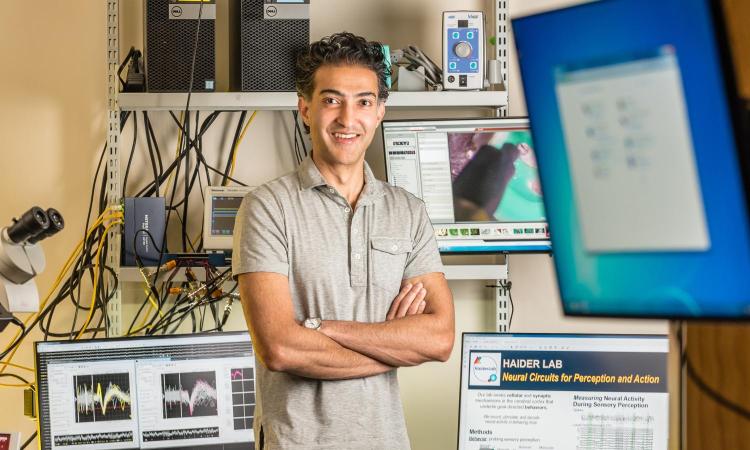 Bilal Haider stands in his lab, with computer monitors in the foreground, a microscope on one side, and more monitors behind him.