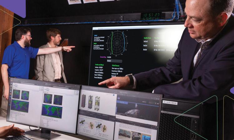 Glenn Lightsey looks over two computer monitors with satellite trajectory information while researchers in the background look at data on a large wall-mounted screen in the background.