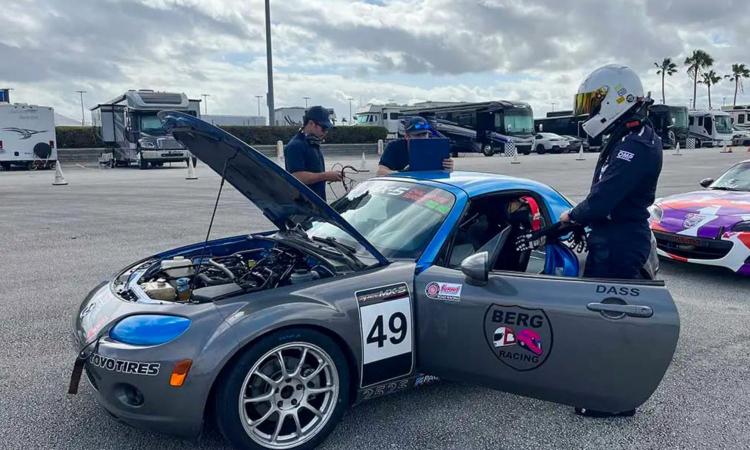 Harbir Dass in racing helmet and jumpsuit preparing to sit in a Mazda MX-5 vehicle with racing decals and its hood raised.