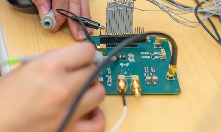 Two hands working on an analog chip (the black TK1 square) on a circuit board that's connected to testing equipment.