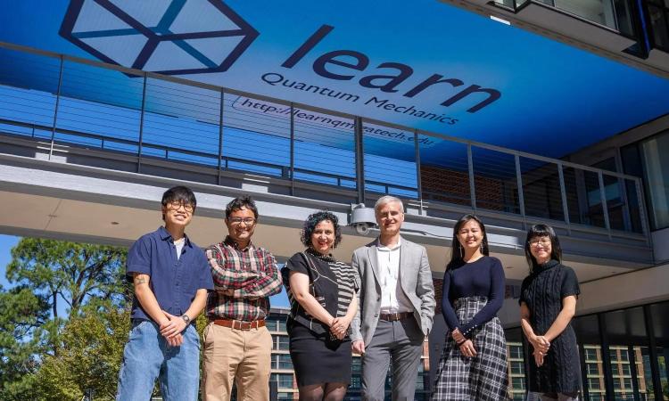 Six members of the research team stand in front of an elevated walkway that has a screen covering its entire ceiling. The screen is displaying a logo and text, "learn quantum mechanics, learnqm.gatech.edu"