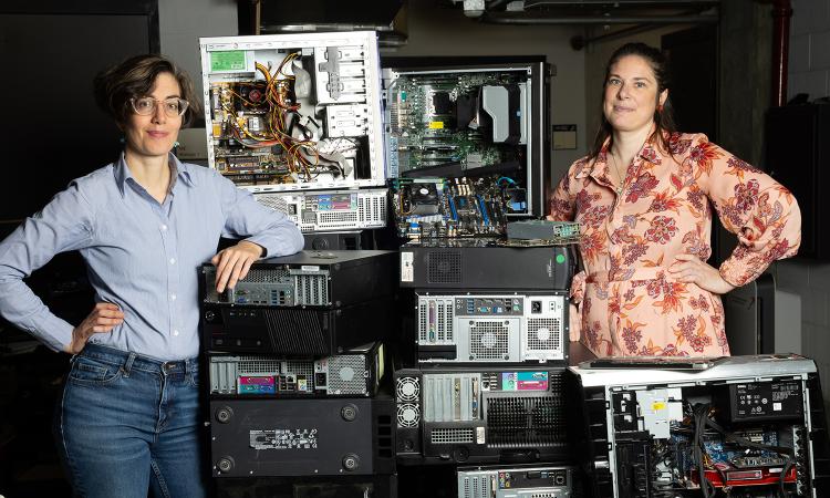 two women stand next to a pile of old computers, circuitboards, and other electronic waste