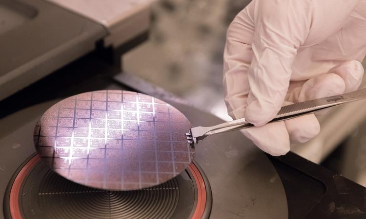 A gloved hand holds uses a metal tool to grasp a round silicon wafer that is reflecting a strip of white light. (Photo: Allison Carter)