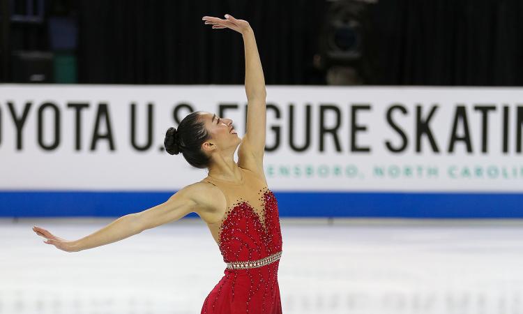 Sierra Venetta in a red dress on the ice during a figure skating routine with her left arm extended upward and her right arm extended behind her. The rink wall in the background reads "Toyota U.S. Figure Skating."
