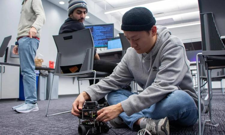 A hackathon participany sits on the floor and works on a small robot while another participant seated at a laptop and monitor with computer code looks on from above.