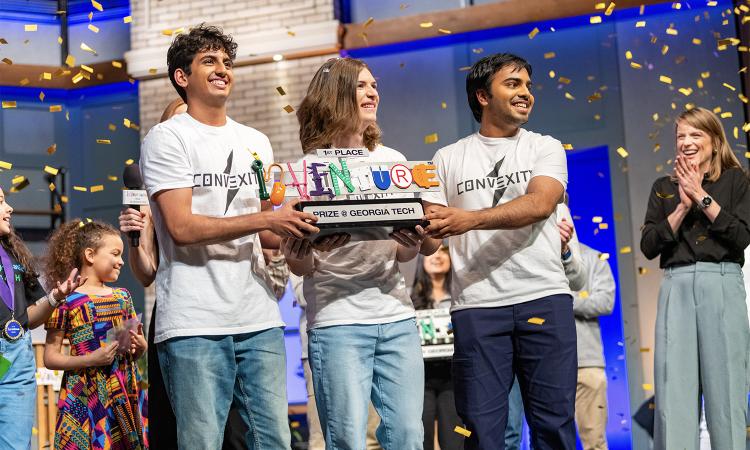 Three people hold a first place trophy with confetti and others in the background