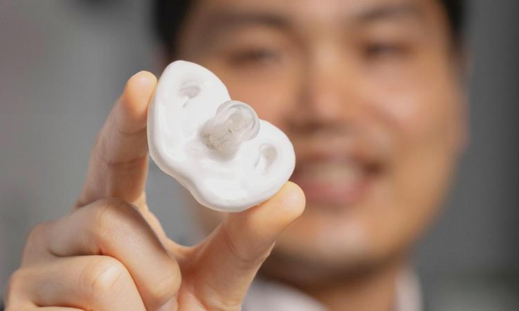 Hong Yeo holds a white prototype electrolyte-sensing pacifier in two fingers. Yeo's face is out of focus in the background.