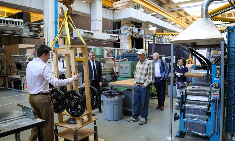 A person stands with hands on a wooden frame with heavy weights suspended from an overhead crane while several other people look on and listen.