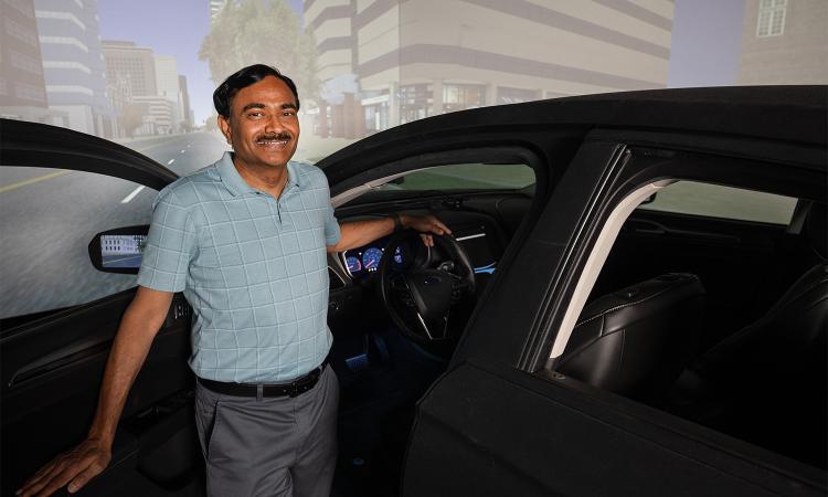 Srinivas Peeta stands next to a car in a simulator lab with a virtual environment on screen behind him.