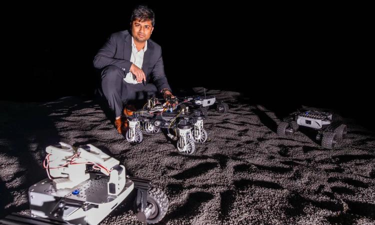 Yashwanth Nakka poses with several rovers in his lab, with simulates the moon's granular terrain and stark lighting of the sun.