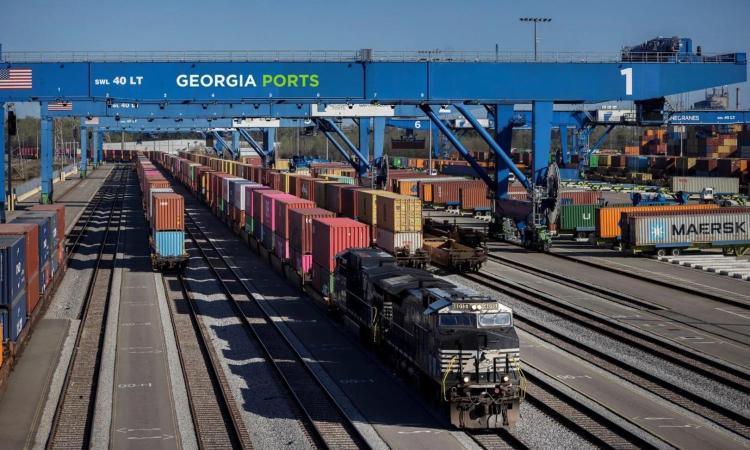 A train pulling a long line of cargo containers in a rail yard under a crane with Georgia Ports on it.