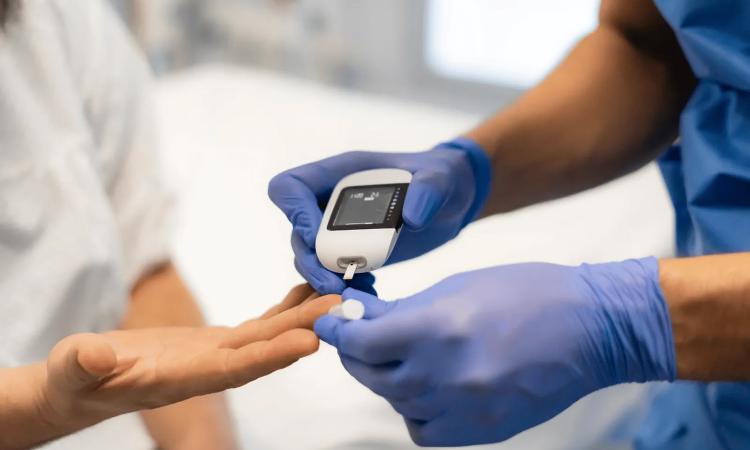 Healthcare worker wearing blue gloves uses a handheld glucose meter to check a patient’s blood sugar from a finger prick.