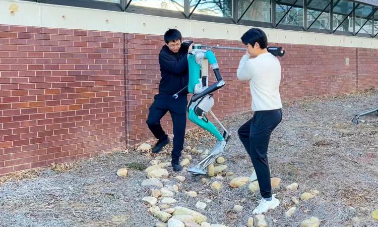 two people with a robot assist it over rocky and uneven terrain beside a building