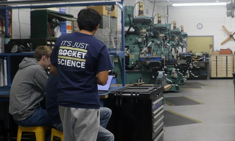 Several students look at a laptop in a machine shop.
