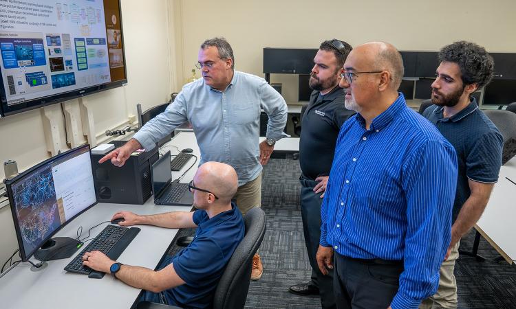 Researchers gathered around a computer and large monitor disucssing their AI model.