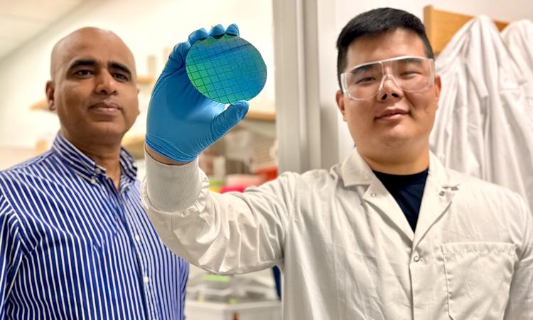 Two researchers in a laboratory examine a blue-green silicon wafer. One, wearing a lab coat, safety goggles, and a blue glove, holds the wafer up for inspection while the other stands beside them. Laboratory equipment is visible in the background.