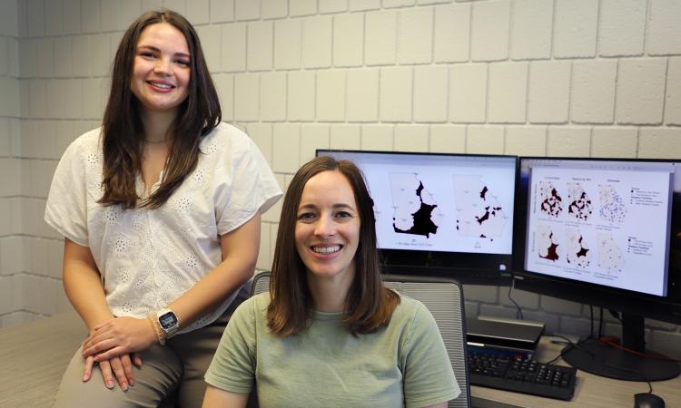Lauren Steimle and Meghan Meredith pose with screens showing data from their maternal healthcare access research.