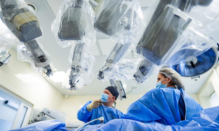 Four robotic arms are poised overhead as a surgeon manipulates a controller in the operating room.
