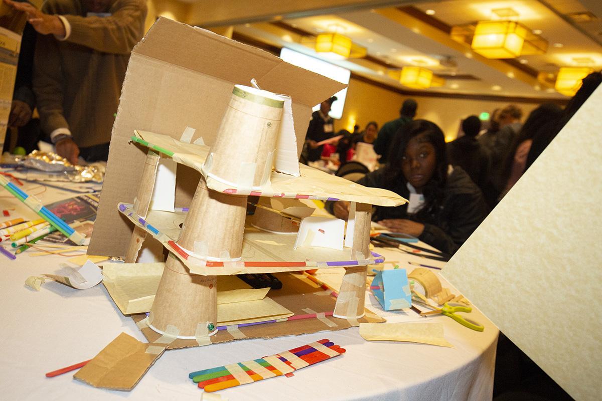 An under-construction three-level cardboard funhouse design with paper cups and straws. The goal was for students to create a path for a golf ball to roll from top to bottom and activate a light or buzzer.