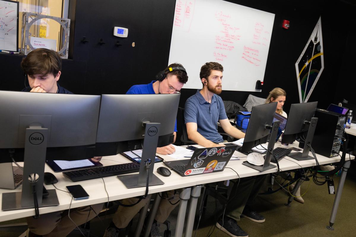 Conner Awald, Mason Starr, and Michael Hauge at their computer terminals in mission control, all looking in different directions. A whiteboard and wall-size logo on are on the black wall behind them.