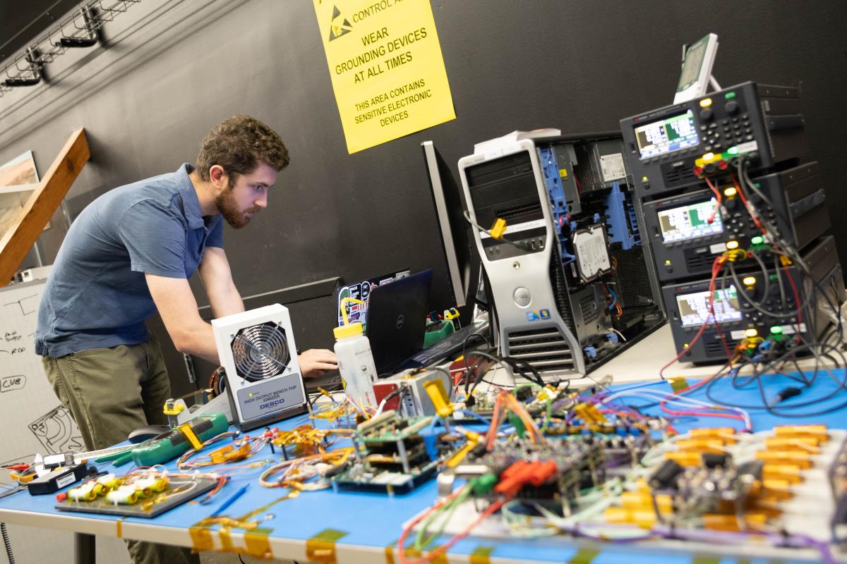 Michael Hauge leans over to work at a computer sitting on a table full of electronic components and wires.