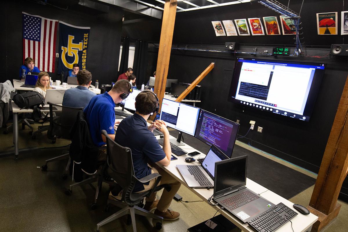 Mission controllers sit at computer screens with a large screen on the opposite wall. In the background, other students sit at a table with computers and USA and Georgia Tech flags hanging on the wall.