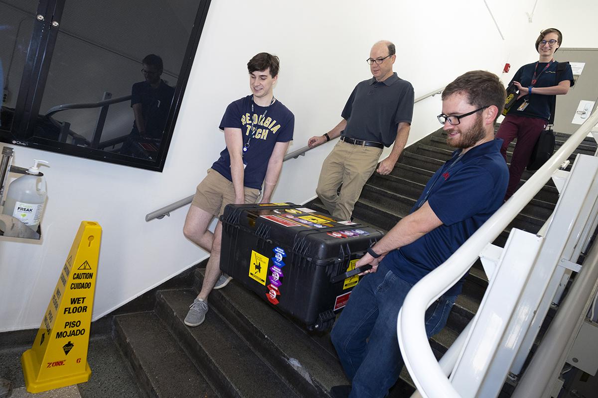 Conner Awald and Nathan Cheek carry a large black case covered with stickers down a staircase. Faculty member Glenn Lightsey and Celeste Smith are walking down behind them.